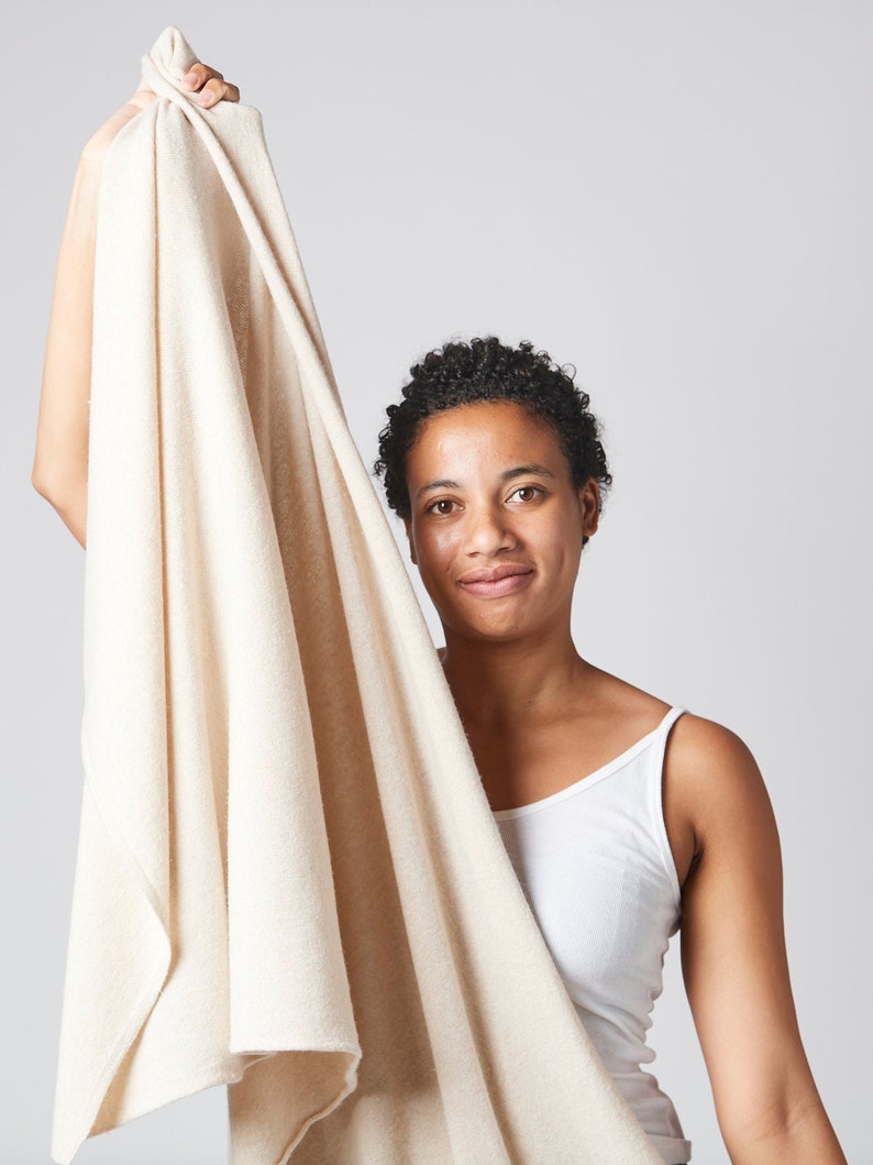 May include: A person holds up a large, off-white towel. The towel is soft and fluffy, and it appears to be made of a natural material. The person is smiling and looking at the camera.