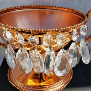 May include: A copper-colored decorative bowl with two handles and a beaded rim. Clear crystal pendants hang from the rim, adding a touch of elegance. The bowl sits on a dark surface.