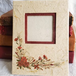 May include: A cream-colored picture frame with a red inner border and a pressed flower design. The frame is made of textured paper and has a rustic look.