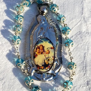 May include: A rosary with light blue beads and a silver-toned pendant. The pendant features an image of a religious scene with angels. The beads are a translucent blue, and the chain is silver.