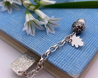May include: A silver bookmark with a detailed design and a chain attached to a silver acorn charm with a leaf. The bookmark is lying on a blue book with white flowers.