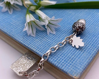 May include: A silver bookmark with an acorn and leaf charm hanging from a chain. The bookmark is lying on a blue book with white flowers.
