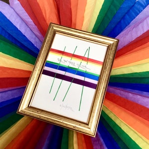 May include: A framed artwork with a gold-colored frame. The artwork features colorful ribbons arranged in a rainbow pattern, with the text "All you need is love." Green lines extend from the ribbons. The background is a rainbow of colorful tissue paper.