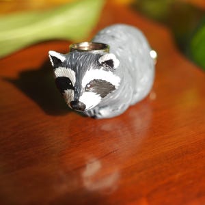 K&ouml;nnte beinhalten: Eine kleine, graue Waschb&auml;r-Figur mit schwarzen Augen und einem wei&szlig;en Streifen im Gesicht. Die Figur tr&auml;gt einen goldenen Ring auf dem Kopf.