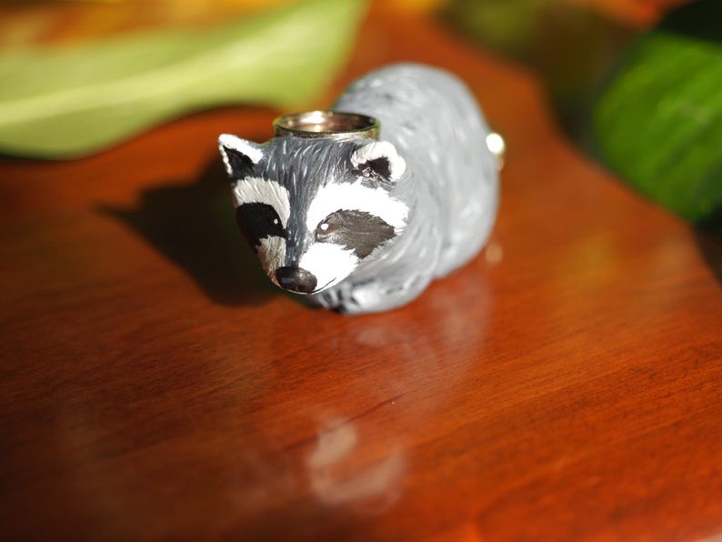 K&ouml;nnte beinhalten: Eine kleine, graue Waschb&auml;r-Figur mit schwarzen Augen und einem wei&szlig;en Streifen im Gesicht. Die Figur tr&auml;gt einen goldenen Ring auf dem Kopf.