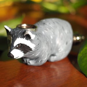 K&ouml;nnte beinhalten: Eine graue Waschb&auml;r-Figur mit schwarzen Augen und einem wei&szlig;en Streifen im Gesicht. Die Figur h&auml;lt einen silbernen Ring im Maul.