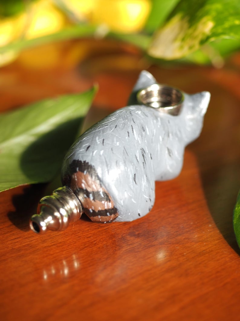 K&ouml;nnte beinhalten: Eine graue Keramik-Waschb&auml;r-Figur mit einem braunen und schwarzen gestreiften Schwanz. Die Figur hat ein kleines Loch oben auf dem Kopf, was darauf hindeutet, dass es sich um eine Pfeife handeln k&ouml;nnte.