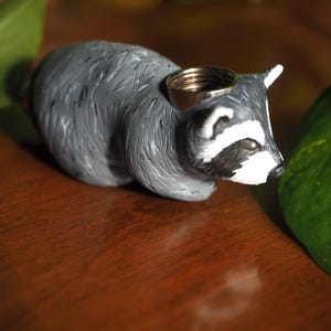 K&ouml;nnte beinhalten: Eine graue und wei&szlig;e Waschb&auml;r-Figur mit schwarzer Nase und Augen. Die Figur hat einen Metallring auf dem Kopf.