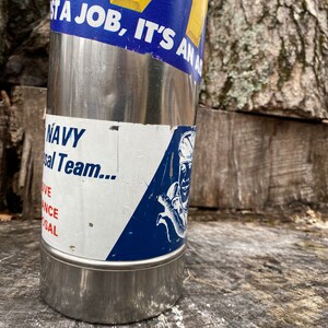 May include: A stainless steel water bottle with a blue and yellow sticker that reads "IT'S AN AD". The bottle also has a sticker that says "NAVY" and features a graphic of a sailor. The bottle is cylindrical and has a shiny, metallic finish.