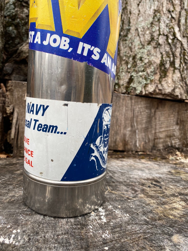 May include: A stainless steel water bottle with a blue and yellow sticker that reads "IT'S AN AD". The bottle also has a sticker that says "NAVY" and features a graphic of a sailor. The bottle is cylindrical and has a shiny, metallic finish.