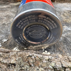 May include: A vintage Aladdin Stanley vacuum bottle. The bottle is silver with a red and blue top. The bottom of the bottle is black with white text that reads "Aladdin Stanley Vacuum Bottle 82". The bottle has instructions printed on it.
