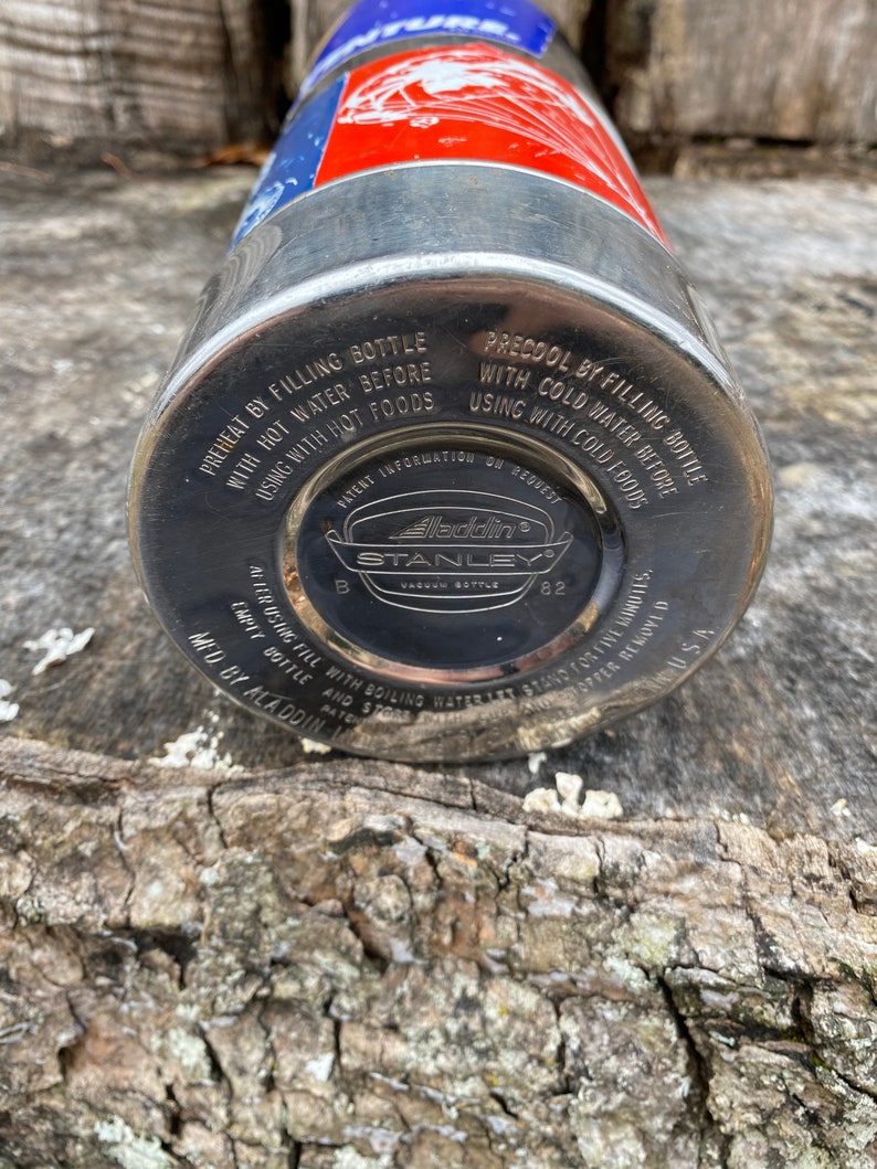 May include: A vintage Aladdin Stanley vacuum bottle. The bottle is silver with a red and blue top. The bottom of the bottle is black with white text that reads "Aladdin Stanley Vacuum Bottle 82". The bottle has instructions printed on it.
