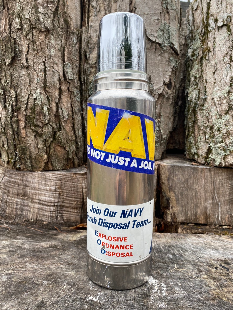 May include: A silver-colored thermos with a shiny, chrome-like lid. The thermos has a blue and yellow sticker that reads "NAVY" and "NOT JUST A JOB." Below, a sticker says "Join Our NAVY Bomb Disposal Team... EXPLOSIVE ORDNANCE DISPOSAL."