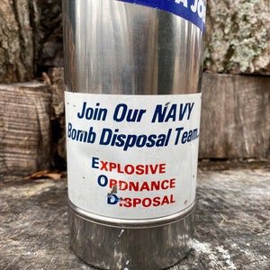 May include: A stainless steel tumbler with a white label featuring the text "Join Our NAVY Bomb Disposal Team" and "EXPLOSIVE ORDNANCE DISPOSAL" in blue and red. The top of the tumbler has a blue and yellow sticker that reads "IT'S NOT JUST A JOB".