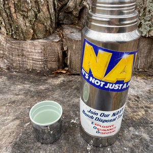 May include: A stainless steel thermos with a light green lid and a matching cup. The thermos has a blue and yellow sticker that says "NAVY" and "IT'S NOT JUST A". A second sticker says "Join Our NAVY Bomb Disposal Team".