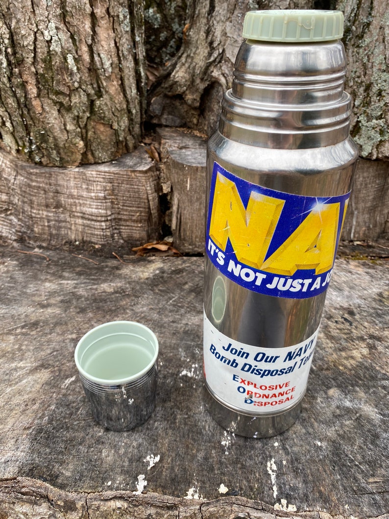 May include: A stainless steel thermos with a light green lid and a matching cup. The thermos has a blue and yellow sticker that says "NAVY" and "IT'S NOT JUST A". A second sticker says "Join Our NAVY Bomb Disposal Team".