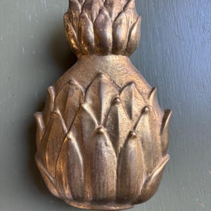 May include: A gold-colored, artichoke-shaped decorative object. The object has a textured surface with detailed leaf-like structures. The artichoke design is a classic decorative element, often used in home decor.