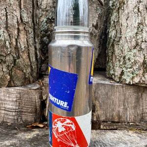 May include: A vintage stainless steel thermos with a clear plastic cup. The thermos has several stickers, including one with the word "ADVENTURE" and another with a parachute design. The thermos is cylindrical and has a silver finish.