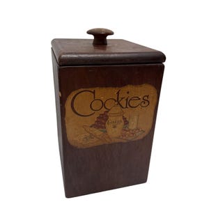 May include: A dark brown wooden cookie jar with a lid and a small knob. The jar features a tan oval-shaped graphic with the word "Cookies" and a baking scene.