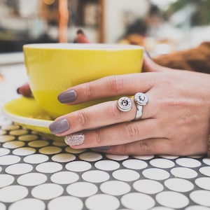 May include: A close-up of a hand wearing a silver ring with two bullet casings. The hand is holding a yellow coffee cup. The hand has gray nail polish and one finger has glitter nail polish.