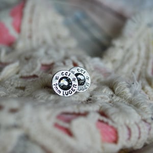 Puede incluir: Un par de pendientes de plata con piedras de cristal negro. Los pendientes tienen forma de casquillos de bala y llevan grabado el texto "CCI 9mm LUGER".