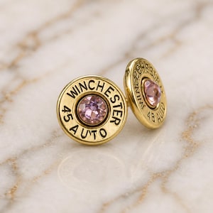 May include: A pair of gold-coloured stud earrings. Each earring is shaped like a bullet casing with the text "WINCHESTER 45 AUTO" engraved around a central pink gemstone. The earrings are set against a marble background.