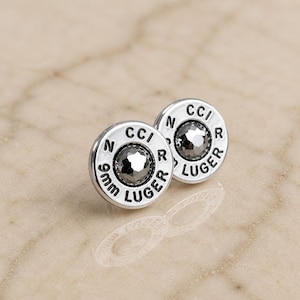 May include: A pair of silver-coloured stud earrings. Each earring is circular with the text "9mm LUGER" and "CCI R" around a central, faceted, grey crystal. The earrings are displayed on a reflective surface.