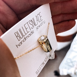 May include: A gold chain necklace with a bullet casing pendant and a clear crystal. The pendant has a small diamond-shaped stone on the top. The necklace is on a white card with the text "BULLETSNLACE handmade jewellery" and "BULLETSNLACE" at the bottom.