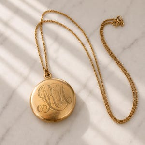 May include: A gold locket necklace with a fine chain. The circular locket has an engraved monogram. The necklace is presented on a white marble surface, with gentle shadows.