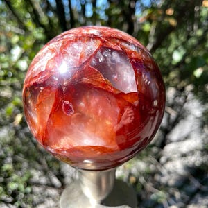 May include: A polished, spherical red and clear crystal stone with internal fractures and deep crimson inclusions, resting on a smooth, metallic silver-toned pedestal stand. The surface of the sphere is reflective and smooth to the touch.