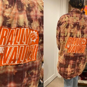 May include: A red and brown plaid flannel shirt with a large orange patch on the back that says "Rally the Valley" with a basketball graphic.