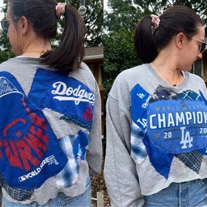 May include: A gray cropped sweatshirt with a patchwork design featuring the Los Angeles Dodgers logo, the words "World Series Champions 2020", and other patches.