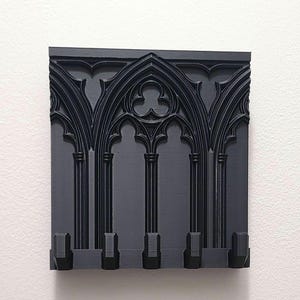 Puede incluir: Un perchero de pared negro de estilo gótico. El perchero presenta un diseño arqueado con columnas verticales y cuatro ganchos en la parte inferior. El diseño recuerda a la arquitectura de las catedrales. El perchero está hecho de un material negro mate sólido.