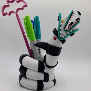 Pode incluir: Um porta-canetas único em forma de verme da areia estilizado, com um corpo listrado em preto e branco e uma cabeça detalhada com dentes afiados. O porta-canetas contém marcadores verdes, azuis e pretos e uma caneta rosa em forma de morcego.