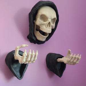Peut inclure: Décoration murale sur le thème d'Halloween. L'ensemble comprend un crâne avec une capuche noire et deux mains squelettiques sortant de manches noires. Le crâne et les mains sont blanc cassé, montés sur un mur violet.