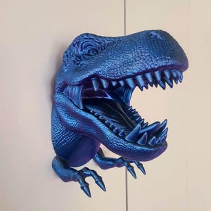 Könnte beinhalten: Eine blaue, metallische Dinosaurierkopfskulptur mit offenem Maul und scharfen Zähnen. Die Skulptur ist an einer Wand befestigt.