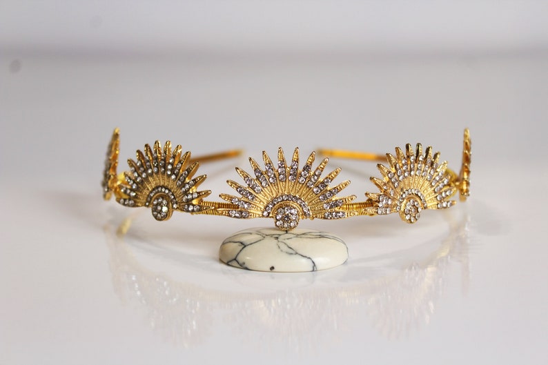 Puede incluir: Una corona dorada con dise&ntilde;o de rayos de sol, adornada con cristales transparentes. La corona se exhibe sobre una piedra blanca y gris, reflejando la luz. Esta joya es adecuada para ocasiones especiales.