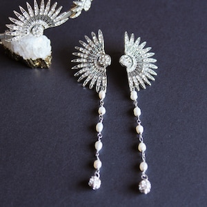 Puede incluir: Pendientes plateados de estilo Art D&eacute;co con un dise&ntilde;o en forma de abanico, adornados con cristales transparentes y hileras de perlas colgantes. Tambi&eacute;n se ve una tiara a juego con adornos similares. Las joyas se exhiben sobre una superficie gris oscuro.