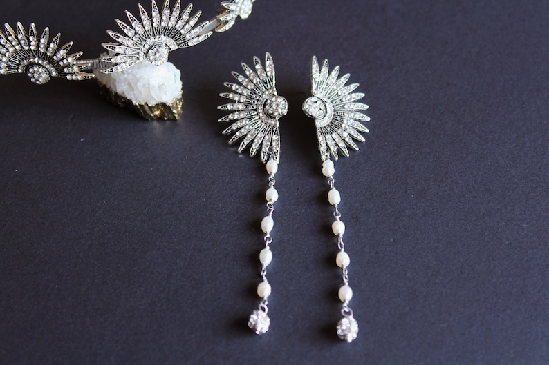 Puede incluir: Pendientes plateados de estilo Art D&eacute;co con un dise&ntilde;o en forma de abanico, adornados con cristales transparentes y hileras de perlas colgantes. Tambi&eacute;n se ve una tiara a juego con adornos similares. Las joyas se exhiben sobre una superficie gris oscuro.