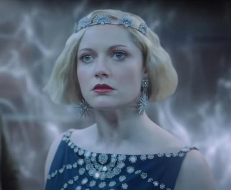 Puede incluir: Una mujer con cabello rubio lleva una tiara plateada y pendientes en forma de estrella. Lleva un vestido azul oscuro con adornos plateados alrededor del escote. La imagen tiene una cualidad suave y et&eacute;rea.