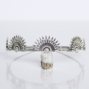 Puede incluir: Corona plateada de estilo Art D&eacute;co con tres adornos en forma de abanico. El adorno central est&aacute; adornado con numerosos cristales transparentes. La corona se muestra sobre una superficie blanca. Tocado decorativo para ocasiones especiales.