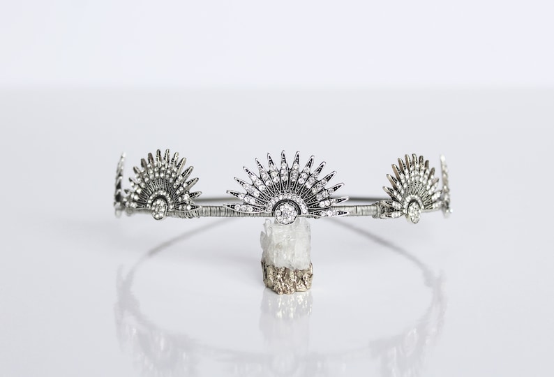 Puede incluir: Corona plateada de estilo Art D&eacute;co con tres adornos en forma de abanico. El adorno central est&aacute; adornado con numerosos cristales transparentes. La corona se muestra sobre una superficie blanca. Tocado decorativo para ocasiones especiales.