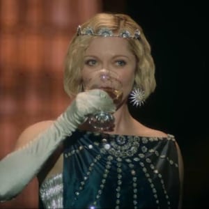 Puede incluir: Una mujer con una tiara enjoyada y un pendiente en forma de estrella bebe de una copa de cristal. Lleva un vestido azul oscuro con adornos plateados y guantes largos blancos. El fondo es borroso y de tonos c&aacute;lidos.