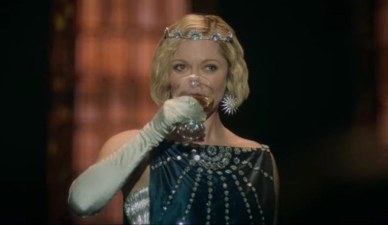 Puede incluir: Una mujer con una tiara enjoyada y un pendiente en forma de estrella bebe de una copa de cristal. Lleva un vestido azul oscuro con adornos plateados y guantes largos blancos. El fondo es borroso y de tonos c&aacute;lidos.