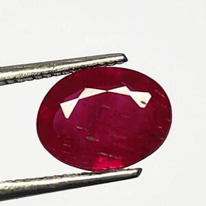 Może przedstawiać: Owalny, żywy czerwony kamień szlachetny trzymany przez pęsetę. Kamień szlachetny ma gładką, wypolerowaną powierzchnię i lekko prześwitujący wygląd.