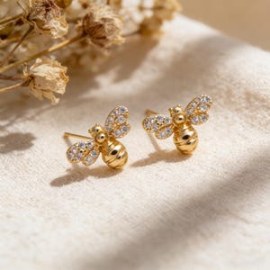 Pode incluir: Um par de brincos de pressão dourados em forma de abelha. Cada abelha tem um corpo listrado e asas adornadas com pequenos cristais transparentes. Os brincos são exibidos em tecido creme, com flores secas ao fundo.