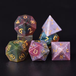 Könnte beinhalten: Ein Satz Edelsteinwürfel in verschiedenen Formen, darunter ein d4, d6, d8, d10, d12 und d20. Die Würfel sind hauptsächlich rot, grün und lila mit goldfarbenen Zahlen. Die Würfel sind auf einer schwarzen Oberfläche angeordnet.