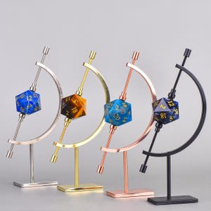 May include: Four dice displays in silver, gold, copper, and black. Each display holds a gemstone D20 die with gold numbers. The dice are blue, brown, and blue-green. The displays have a curved arm and a base.
