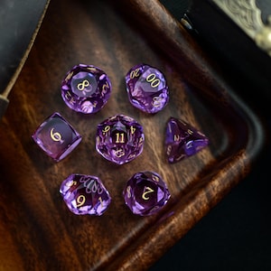 Op de afbeelding: Set van zeven paarse en gouden dobbelstenen met verschillende nummers op elke zijde. De dobbelstenen bevinden zich in een houten bak.