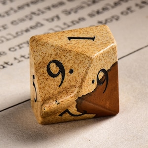 May include: A six-sided die with a textured, tan surface and dark brown accents. The die features black numerals, including a "1" and multiple "9"s. The die is resting on a surface with faint text.